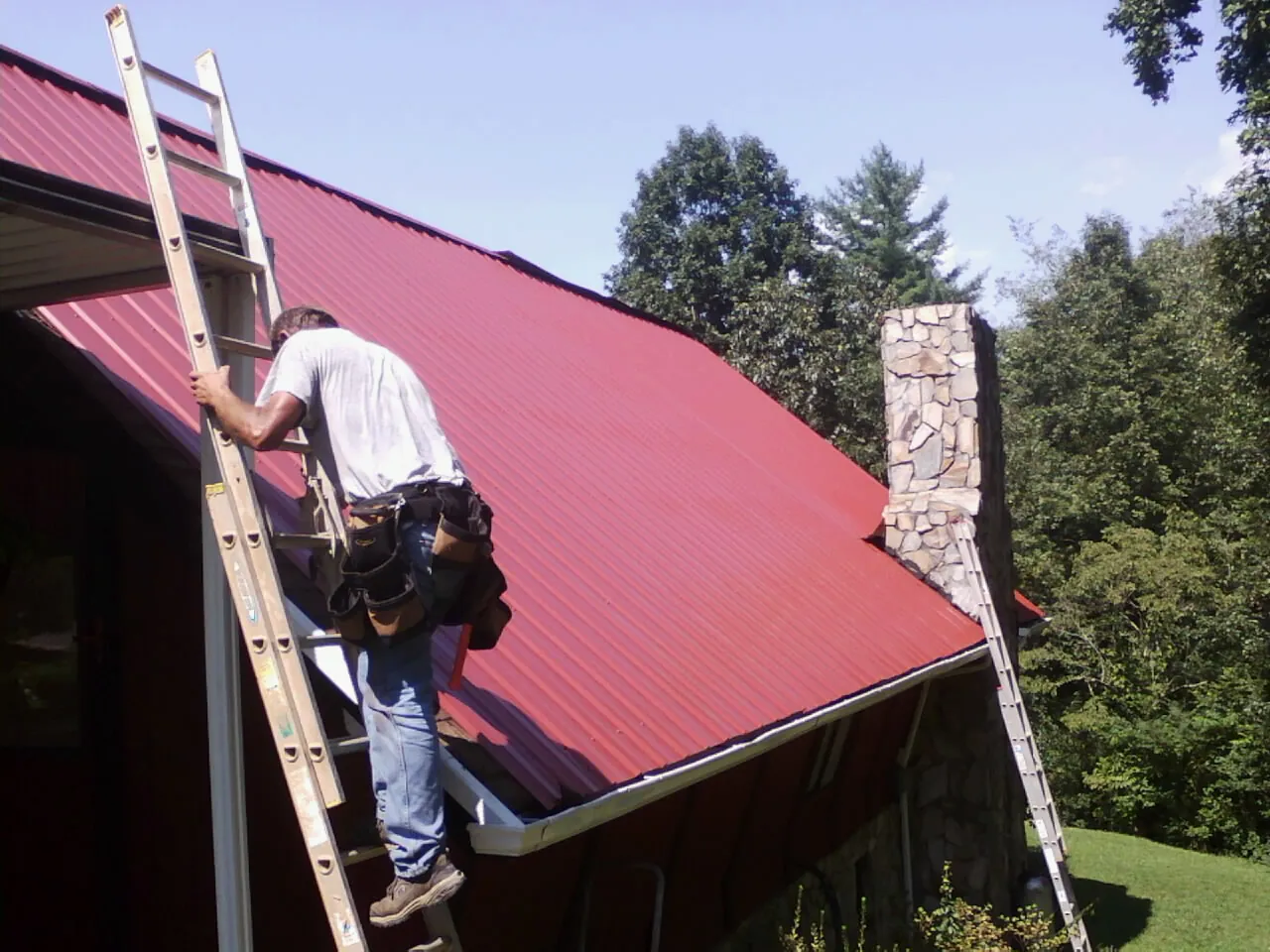 Roofer on a ladder beside a red metal roof and stone chimney — Soffit & Fascia Repair by Roofing Brothers in Carbon Hill